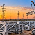 Skipper: steel transmission tower sections stacked in a yard at sunset as a crane lifts one, with pylons behind