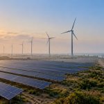 Wind turbines behind rows of solar panels on a coastal plain at sunrise with a power line along a dirt road