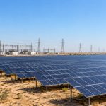Rows of solar panels on dry land in Rajasthan with a substation and transmission towers in the background