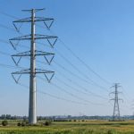 A tall four-circuit pole-type transmission structure with power lines across farmland and lattice towers in the distance