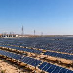 Rows of solar panels on open desert land with a substation and transmission towers in the distance